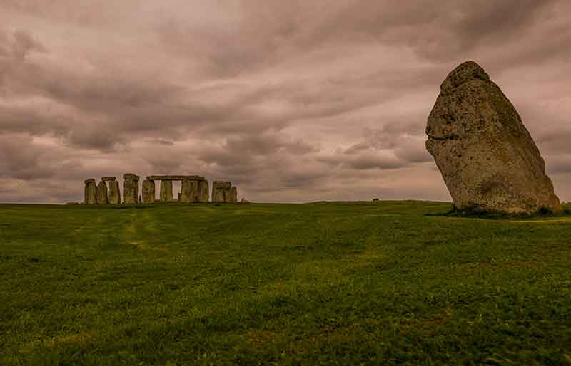 Heel stone in foreground with the main circle behind beneath a dramatic cloudy sky.